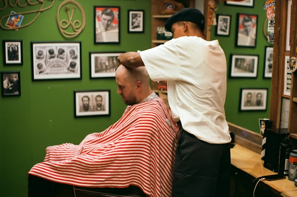 A barber wearing a cap is giving a haircut to a customer draped in a red and white striped cape. The setting is a barbershop with green walls adorned with various framed photographs and memorabilia.