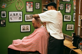 A barber wearing a cap is giving a haircut to a customer draped in a red and white striped cape. The setting is a barbershop with green walls adorned with various framed photographs and memorabilia.