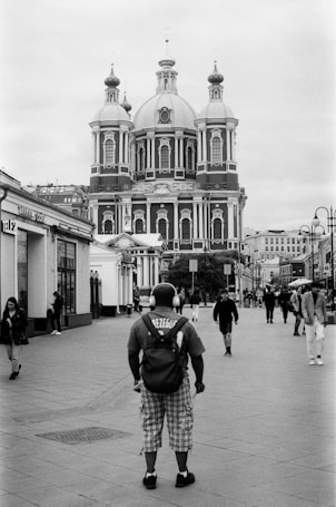 A black and white photograph featuring a large, ornate building with multiple domes in the background, likely a church or cathedral. In the foreground, a person wearing headphones and a shirt with text on the back stands on a cobblestone street, with several other pedestrians walking nearby. The scene appears to be in an urban setting with additional buildings visible alongside the street.