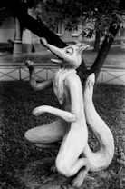 A whimsical sculpture of a cartoonish, anthropomorphic fox in a sitting pose on a grassy area. The fox is designed with exaggerated features such as a long snout and large ears, and its posture suggests a playful or joyful demeanor. The background includes trees and parts of a building, hinting at a park or garden setting.