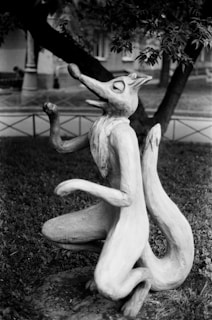A whimsical sculpture of a cartoonish, anthropomorphic fox in a sitting pose on a grassy area. The fox is designed with exaggerated features such as a long snout and large ears, and its posture suggests a playful or joyful demeanor. The background includes trees and parts of a building, hinting at a park or garden setting.
