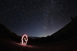A dark night sky is filled with numerous stars, creating a vivid display of the cosmos above a silhouette of a mountainous landscape. In the foreground, a figure is outlined with red light, suggesting the use of long exposure photography to create a glowing effect around the person.