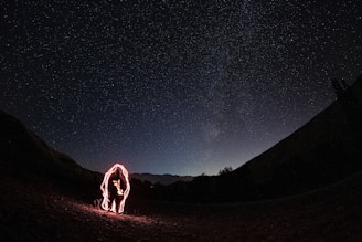 A dark night sky is filled with numerous stars, creating a vivid display of the cosmos above a silhouette of a mountainous landscape. In the foreground, a figure is outlined with red light, suggesting the use of long exposure photography to create a glowing effect around the person.