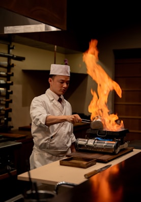 A chef in a white uniform and hat is carefully pouring liquid into a pan, causing a large flame to rise. The kitchen setting appears to be traditional with warm lighting, and there are various cooking utensils and equipment visible.