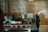 A dimly lit industrial-style kitchen with a vintage boombox and various jars on a shelf. There is a colorful graffiti-style art piece on the wall behind the shelf. A person with a white cap is partially visible, standing next to the shelf.