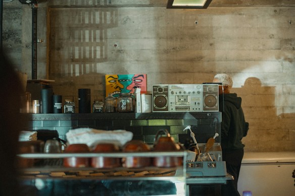 A dimly lit industrial-style kitchen with a vintage boombox and various jars on a shelf. There is a colorful graffiti-style art piece on the wall behind the shelf. A person with a white cap is partially visible, standing next to the shelf.