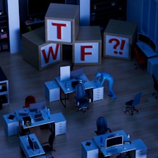 A dimly lit office space with several desks equipped with computers and office chairs. A mannequin-like figure is interacting with one of the desks. Large wooden blocks with the letters 'T', 'W', 'F', and a '?!' symbol are stacked in the background.