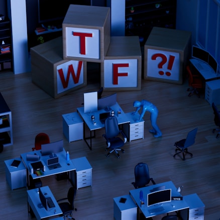 A dimly lit office space with several desks equipped with computers and office chairs. A mannequin-like figure is interacting with one of the desks. Large wooden blocks with the letters 'T', 'W', 'F', and a '?!' symbol are stacked in the background.