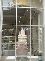 An elaborate multi-tiered cake is displayed in a shop window. The cake features detailed decorations, including patterns and embellishments in pastel colors, and is topped with a pink layer with chocolate drizzle and a decorative cupcake. The shop interior is visible, showcasing shelves with other baked goods.