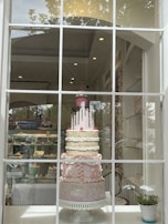 An elaborate multi-tiered cake is displayed in a shop window. The cake features detailed decorations, including patterns and embellishments in pastel colors, and is topped with a pink layer with chocolate drizzle and a decorative cupcake. The shop interior is visible, showcasing shelves with other baked goods.