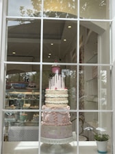 An elaborate multi-tiered cake is displayed in a shop window. The cake features detailed decorations, including patterns and embellishments in pastel colors, and is topped with a pink layer with chocolate drizzle and a decorative cupcake. The shop interior is visible, showcasing shelves with other baked goods.