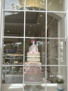 An elaborate multi-tiered cake is displayed in a shop window. The cake features detailed decorations, including patterns and embellishments in pastel colors, and is topped with a pink layer with chocolate drizzle and a decorative cupcake. The shop interior is visible, showcasing shelves with other baked goods.
