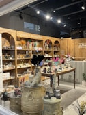 A cozy shop interior displays a variety of candles and accessories. Wooden shelves line the walls, filled with candles, vases, and decorative items. The central wooden table is adorned with an array of colorful dried flowers and various candle options. Large decorative jars and small potted plants add to the rustic aesthetic.