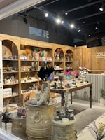 A cozy shop interior displays a variety of candles and accessories. Wooden shelves line the walls, filled with candles, vases, and decorative items. The central wooden table is adorned with an array of colorful dried flowers and various candle options. Large decorative jars and small potted plants add to the rustic aesthetic.