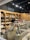 A cozy shop interior displays a variety of candles and accessories. Wooden shelves line the walls, filled with candles, vases, and decorative items. The central wooden table is adorned with an array of colorful dried flowers and various candle options. Large decorative jars and small potted plants add to the rustic aesthetic.
