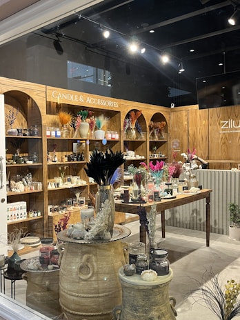 A cozy shop interior displays a variety of candles and accessories. Wooden shelves line the walls, filled with candles, vases, and decorative items. The central wooden table is adorned with an array of colorful dried flowers and various candle options. Large decorative jars and small potted plants add to the rustic aesthetic.