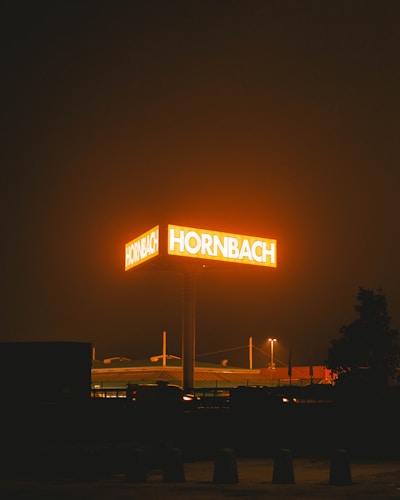A brightly illuminated sign glows with the text 'HORNBACH' against a dark night sky. The orange glow from the sign bathes the surrounding area in a warm light. In the background, the roof of a building and some dimly lit streetlights are visible. The overall scene suggests a commercial area after dark.