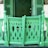 A vibrant green wooden porch railing with ornate cutouts resembling stylized birds or plant shapes. The design features curved patterns and vertical posts, creating an intricate and decorative barrier. In the background, a matching green door is partially visible alongside shutters, contributing to a cohesive color scheme.