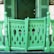 A vibrant green wooden porch railing with ornate cutouts resembling stylized birds or plant shapes. The design features curved patterns and vertical posts, creating an intricate and decorative barrier. In the background, a matching green door is partially visible alongside shutters, contributing to a cohesive color scheme.