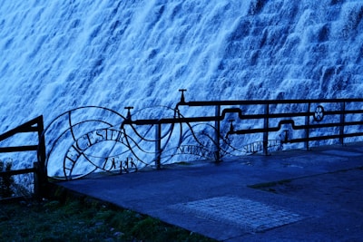 A strong flow of water pours intensely down a sloped concrete surface, resembling a dam or weir structure. A metal railing in the foreground features ornate designs and names of cities, providing a decorative contrast to the turbulent water in the background.