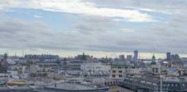 A panoramic view of a cityscape featuring numerous rooftops and a complex skyline. Many buildings have distinct architectural styles with varying roof designs. In the distance, tall modern skyscrapers contrast with historic structures. The sky is cloudy with patches of blue peeking through, suggesting a somewhat overcast weather.