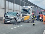 A collision involving a large truck, a van with significant front-end damage, and a street sweeper on a highway. Emergency responders, including firefighters and a person in orange, are present at the scene. The road is partially blocked, and debris is scattered across the pavement.