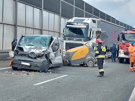 A collision involving a large truck, a van with significant front-end damage, and a street sweeper on a highway. Emergency responders, including firefighters and a person in orange, are present at the scene. The road is partially blocked, and debris is scattered across the pavement.