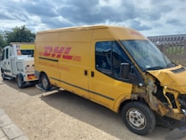 A yellow delivery van with visible front-end damage is being towed by a recovery vehicle. The van belongs to a well-known courier company, as indicated by the branding on its side. The surrounding environment includes a gravel area, a concrete boundary wall, and a partly cloudy sky.