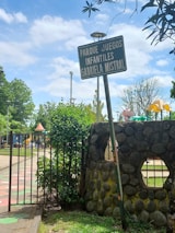 A playground entrance sign labeled 'Parque Juegos Infantiles Gabriela Mistral' stands beside a stone wall and an open gate. Trees and playground equipment are visible in the background, under a partly cloudy sky.