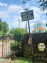 A playground entrance sign labeled 'Parque Juegos Infantiles Gabriela Mistral' stands beside a stone wall and an open gate. Trees and playground equipment are visible in the background, under a partly cloudy sky.