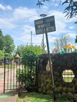 A playground entrance sign labeled 'Parque Juegos Infantiles Gabriela Mistral' stands beside a stone wall and an open gate. Trees and playground equipment are visible in the background, under a partly cloudy sky.