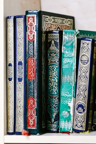 A row of intricately designed, colorful books with ornate calligraphy and decorative patterns. The books are primarily in shades of blue, green, and red, and are arranged neatly on a shelf.
