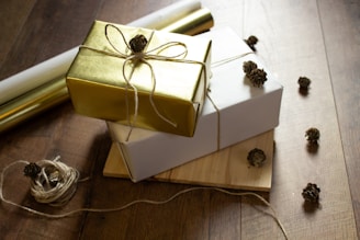 A collection of wrapped gifts arranged on a wooden floor, featuring a gold and white package each tied with twine and adorned with small pinecone decorations. Surrounding the gifts are loose pieces of twine and scattered pinecones, adding a rustic touch.