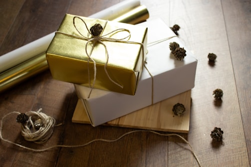 A collection of wrapped gifts arranged on a wooden floor, featuring a gold and white package each tied with twine and adorned with small pinecone decorations. Surrounding the gifts are loose pieces of twine and scattered pinecones, adding a rustic touch.