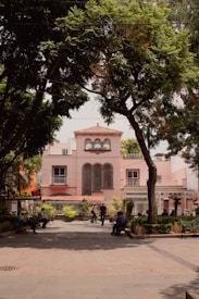 A large pink building with arched windows stands prominently in the background, partially framed by lush green trees. Several people are walking or sitting on benches in a plaza area in front of the building. A person is seen with a bicycle, and there is a sign with the text 'CASA DE ARTESANO' visible on the left.