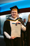 A person wearing a graduation cap and gown is holding a certificate, standing indoors. The background features people and a wall with blue lighting.