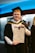 A person wearing a graduation cap and gown is holding a certificate, standing indoors. The background features people and a wall with blue lighting.