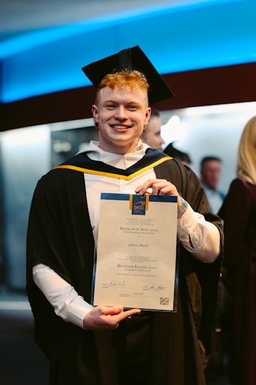 A person wearing a graduation cap and gown is holding a certificate, standing indoors. The background features people and a wall with blue lighting.