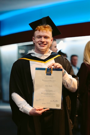 A person wearing a graduation cap and gown is holding a certificate, standing indoors. The background features people and a wall with blue lighting.