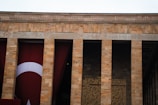 A stone building facade with tall pillars and a large Turkish flag. The architecture features vertical columns with inscriptions visible between them.