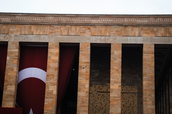 A stone building facade with tall pillars and a large Turkish flag. The architecture features vertical columns with inscriptions visible between them.