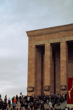 A large crowd gathers at the base of a monumental stone building with tall columns. People are holding flags, and there are floral wreaths mounted on the structure. The sky is overcast, adding to the solemn atmosphere.