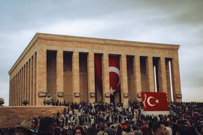 A large, neoclassical building with tall columns and flags featuring the Turkish crescent and star. A large crowd of people is gathered in front of the structure, some holding smaller flags. The sky is overcast, lending a somber mood to the scene.