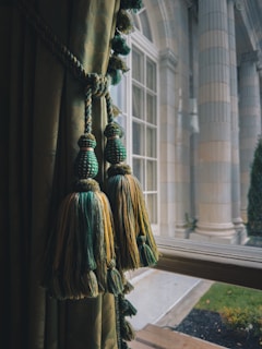 Elegant window treatments with green and gold tassels hang beside a classic, large glass window. The view outside reveals grand architectural columns and a manicured garden area, indicating a sophisticated setting.