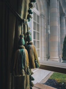 Elegant window treatments with green and gold tassels hang beside a classic, large glass window. The view outside reveals grand architectural columns and a manicured garden area, indicating a sophisticated setting.