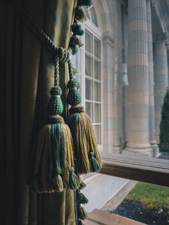 Elegant window treatments with green and gold tassels hang beside a classic, large glass window. The view outside reveals grand architectural columns and a manicured garden area, indicating a sophisticated setting.