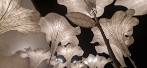 Large, elegant flower-shaped lamps made of translucent material are illuminated against a dark background. The design features organic curves and lines, resembling petals unfolding towards the sky. The lighting creates a soft, atmospheric glow that enhances the texture and structure of the lamps.