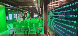A modern room with green walls and chairs, featuring digital screens and displays. There are rows of green chairs facing a main screen at the front of the room, with people seated and a few standing. The room has a high-tech atmosphere, with a large wall display of green binary code on one side.
