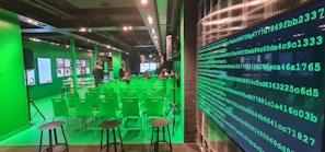 A modern room with green walls and chairs, featuring digital screens and displays. There are rows of green chairs facing a main screen at the front of the room, with people seated and a few standing. The room has a high-tech atmosphere, with a large wall display of green binary code on one side.
