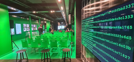 A modern room with green walls and chairs, featuring digital screens and displays. There are rows of green chairs facing a main screen at the front of the room, with people seated and a few standing. The room has a high-tech atmosphere, with a large wall display of green binary code on one side.